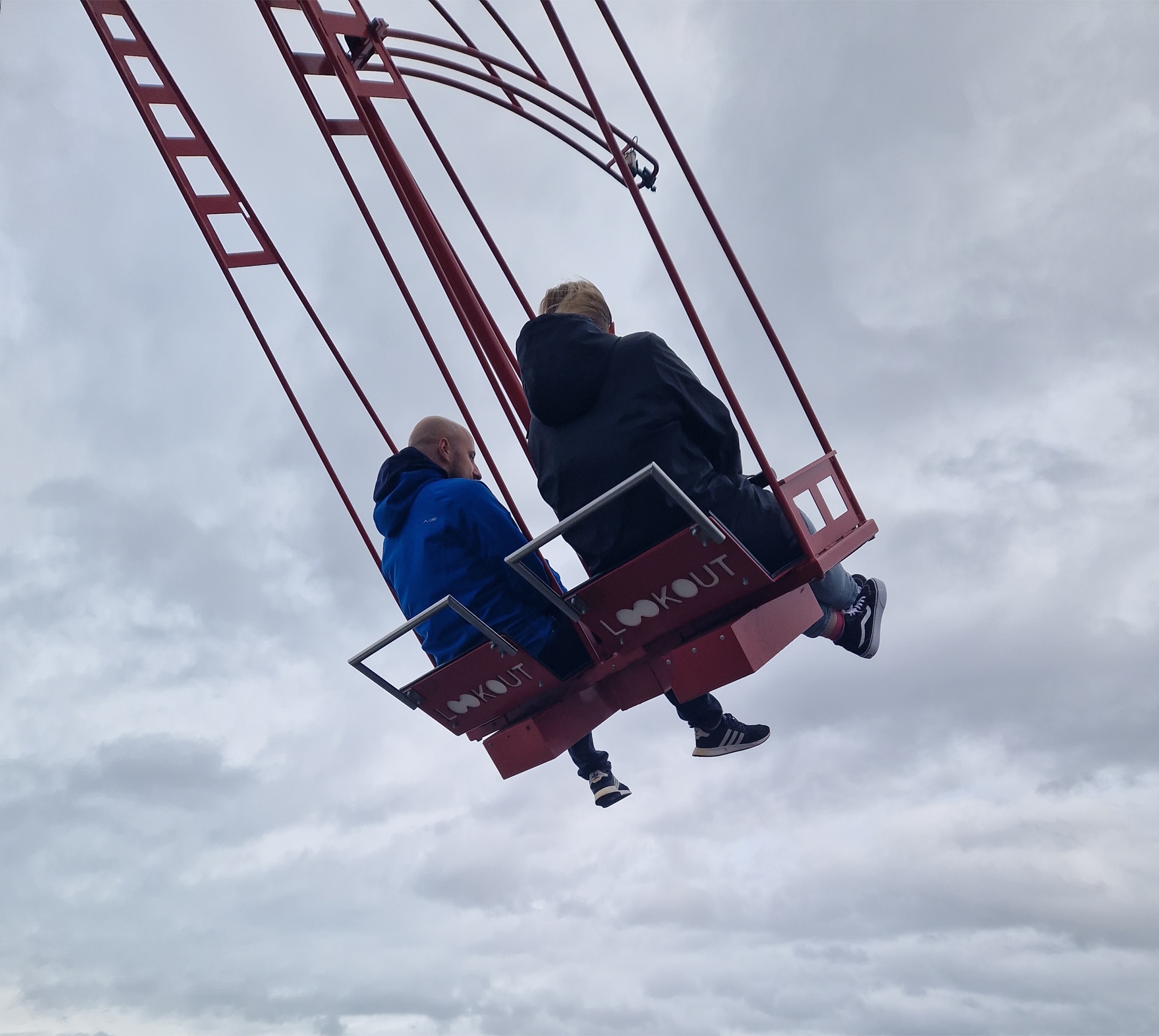 Two riders swung out over the IJ on the Over The Edge swing, seen from further back with the whole rig against an overcast sky